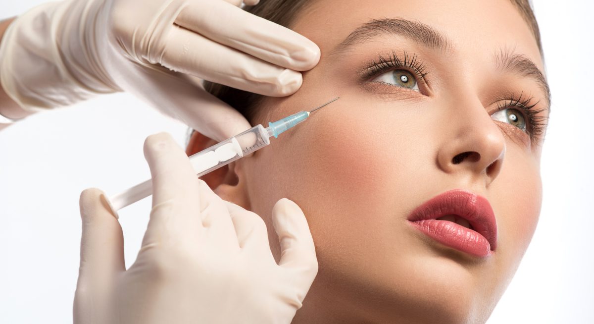 Cute girl receiving hyaluronic acid treatment Serine young woman is getting facial botox injection. Beautician hands in gloves holding syringe near her face